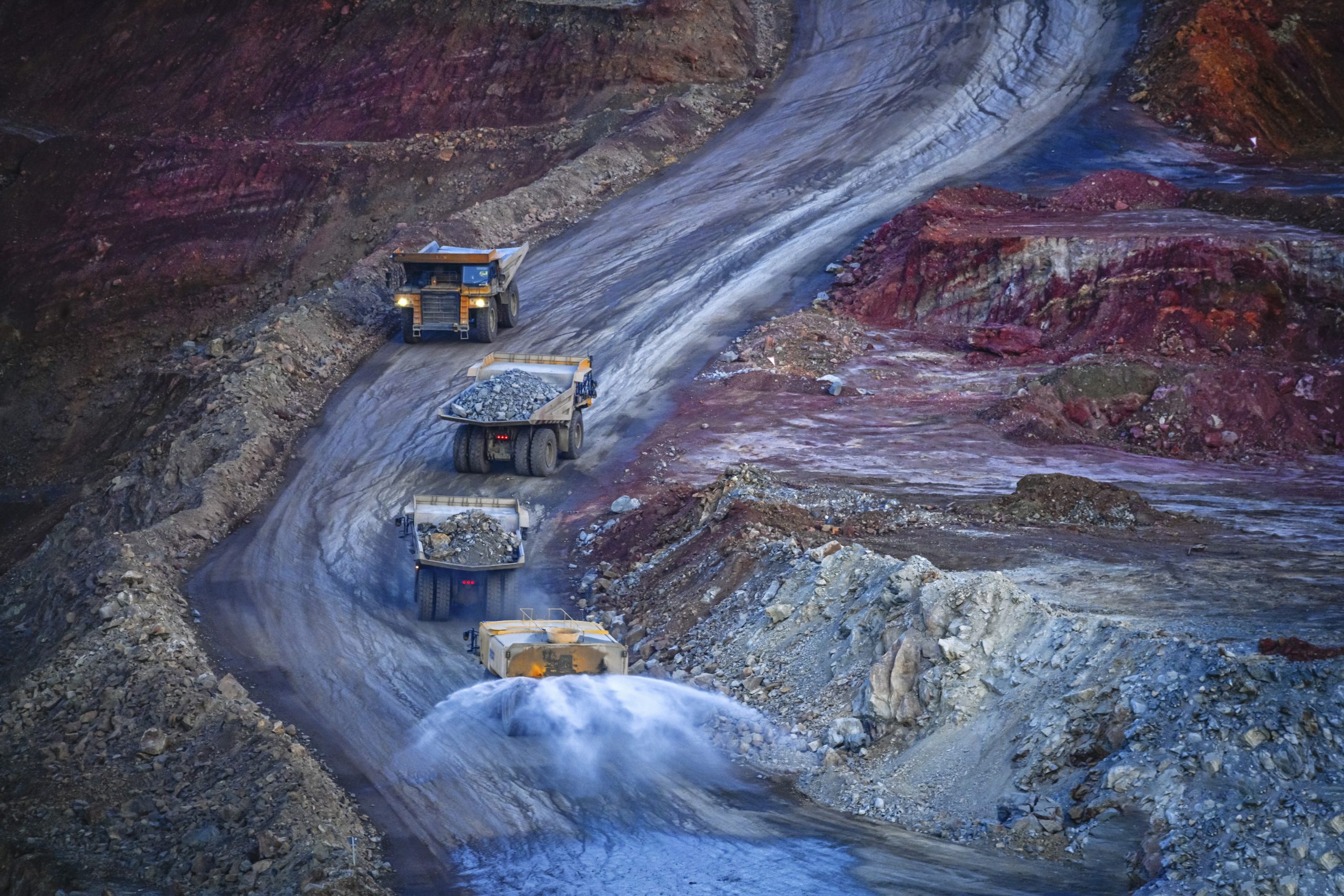 Heavy mining trucks navigate the winding terraced road of a deep pit mine, with exposed mineral strata visible