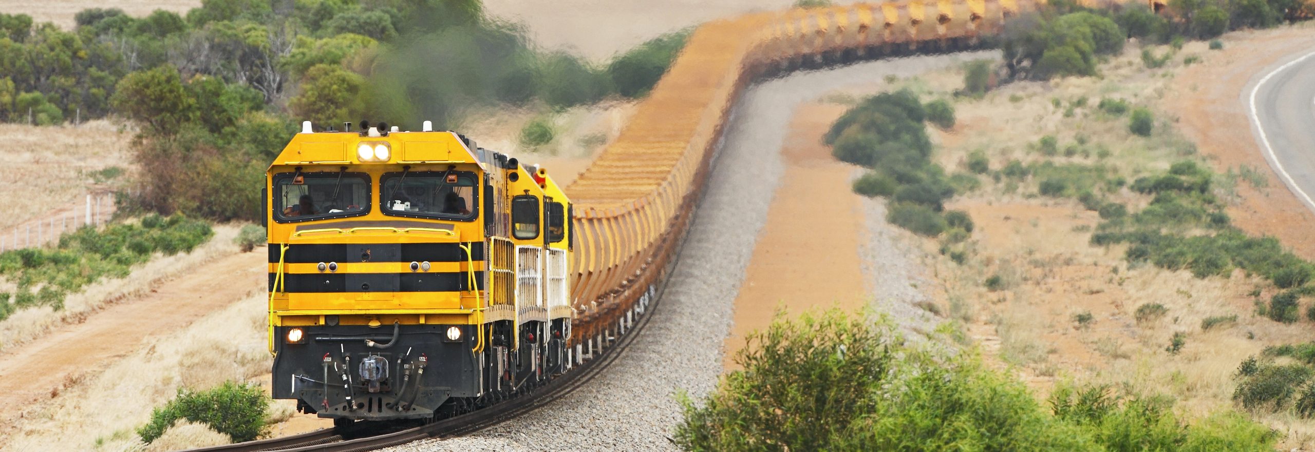 Empty iron ore train with bright orange freight wagons (cars) passes through rolling hills.  Horizontal, telephoto, panorama format, ID removed, corporate colours changed, copy space.